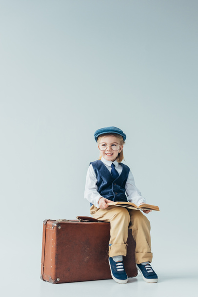 smiling kid in retro vest and cap sitting on suitcase and holding book on grey background - Photo, Image