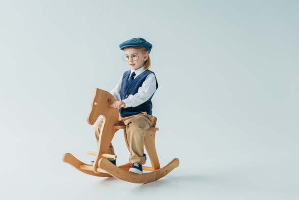 Kind in Retro-Weste und Mütze reitet auf Schaukelpferd und schaut weg  - Foto, Bild
