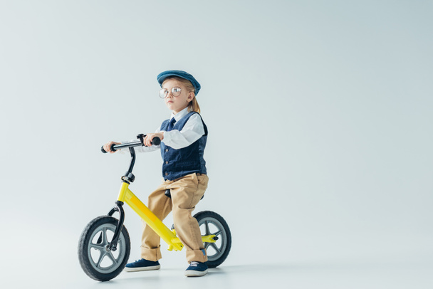вдумчивый ребенок в ретро жилете и кепке катается на велосипеде и смотрит в сторону
  - Фото, изображение