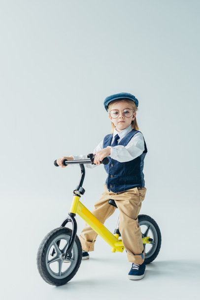 serious kid in retro vest and cap riding bike and looking at camera on grey background - Fotoğraf, Görsel