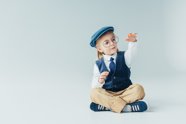 adorable kid sitting on floor with crossed legs and holding butterfly on grey background  - Photo, Image