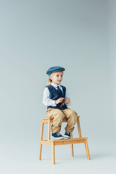 adorable kid sitting on wooden stairs, holding glasses and looking away on grey background  - Photo, Image