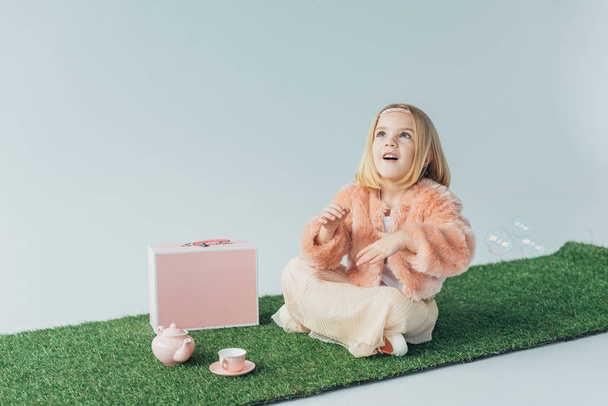 cute kid with crossed legs sitting on grass rug and looking at soap bubbles isolated on grey - Fotoğraf, Görsel