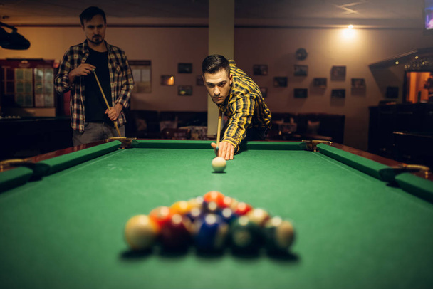 Two male billiard players with cue at the table with colorful balls. Men plays american pool in sport bar - Fotó, kép