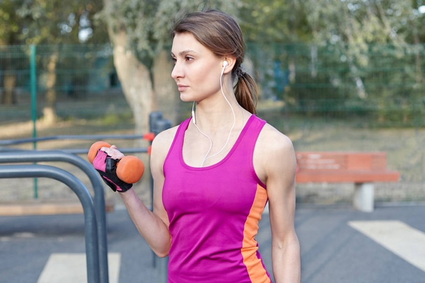 Beautiful slim and sporty girl in bright sportswear trains with dumbbell for biceps at outdoor sportground. White earphones, protective gloves. Real woman in morning training, strenght and motivation. Copy space - Foto, afbeelding