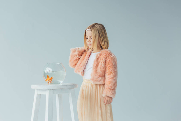 cute kid in faux fur coat and skirt looking at fishbowl on stool isolated on grey - Foto, afbeelding