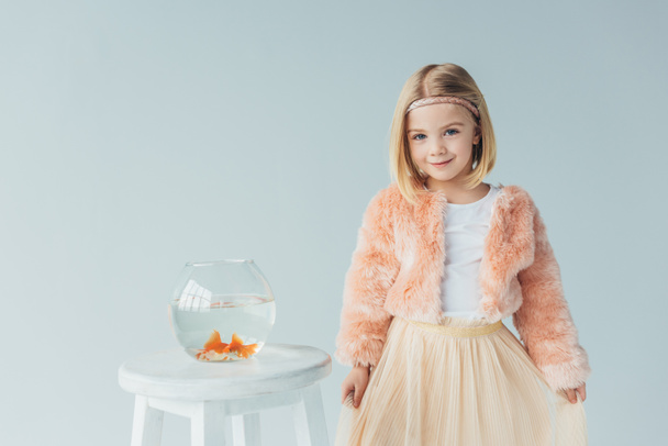 adorable kid in faux fur coat and skirt looking at camera and standing near to stool with fishbowl isolated on grey - Фото, зображення
