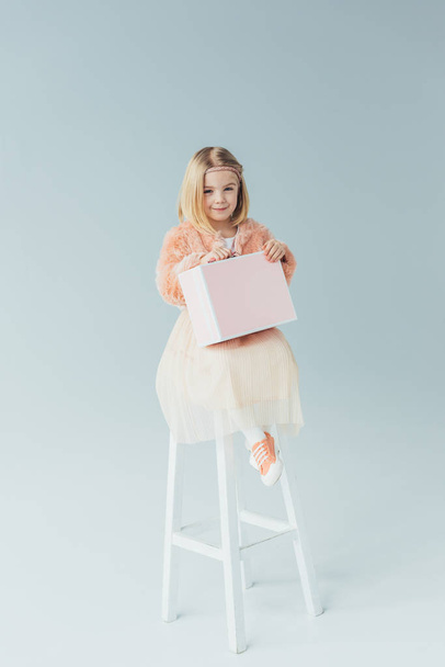 kid in faux fur coat and skirt sitting on highchair and holding pink case on grey background - Photo, image