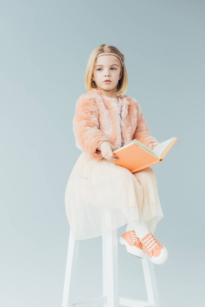  cute kid in faux fur coat and skirt sitting on highchair and holding book isolated on grey  - Photo, Image