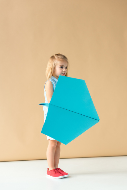 adorable kid playing with blue paper plane on beige background  - Fotografie, Obrázek