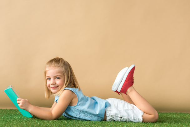 bambino sorridente e carino sdraiato sul tappeto di erba e tenendo libro su sfondo beige
  - Foto, immagini