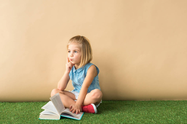  cute child sitting with crossed legs on grass rug and holding book on beige background  - Photo, Image
