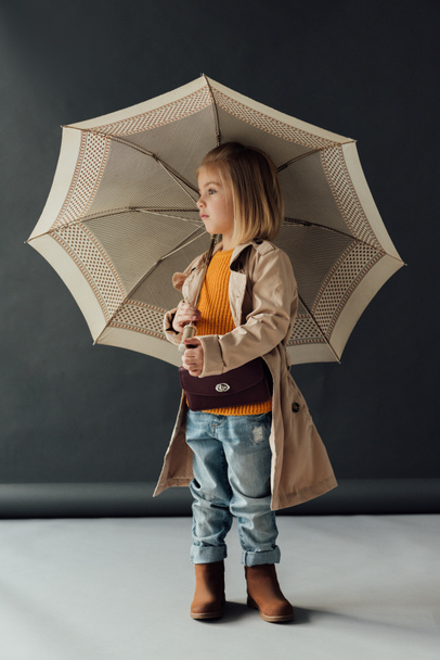 serious child in trench coat and jeans holding umbrella and looking away  - Foto, Bild