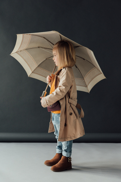side view of child in trench coat and jeans holding umbrella on black background - Photo, Image