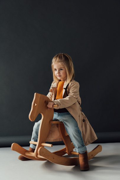  child in trench coat and jeans sitting on rocking horse on black background - Foto, Imagem