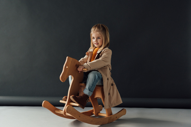  smiling child in trench coat and jeans sitting on rocking horse on black background - Photo, Image