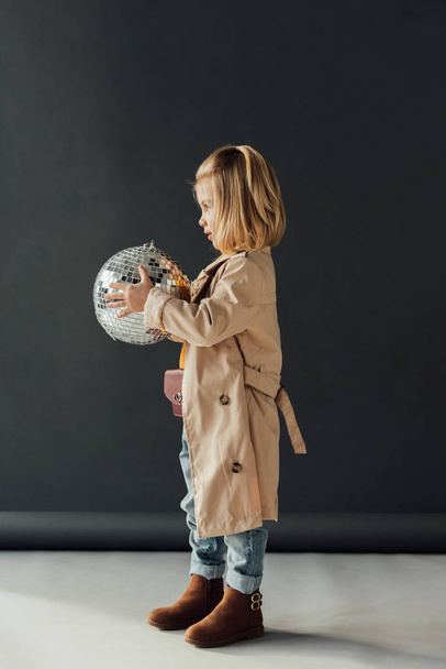 side view of child in trench coat holding disco ball on black background - Φωτογραφία, εικόνα