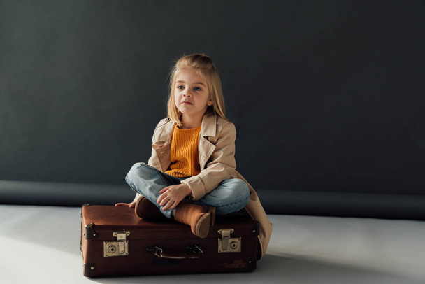 pensive child sitting with crossed legs on leather suitcase on black background - Φωτογραφία, εικόνα