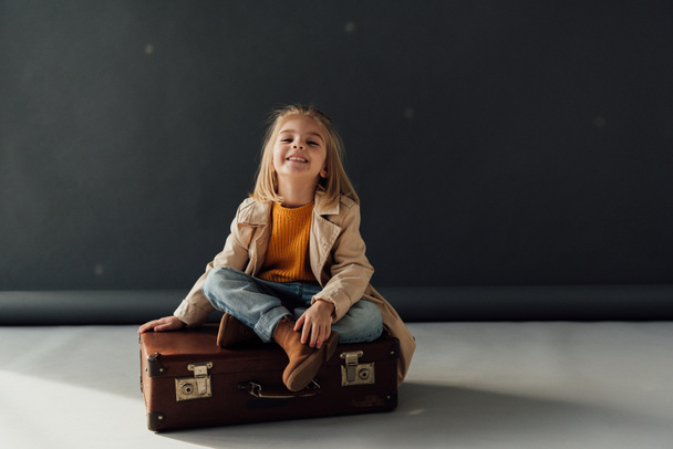smiling and cute child sitting with crossed legs on leather suitcase on black background - Foto, Imagen