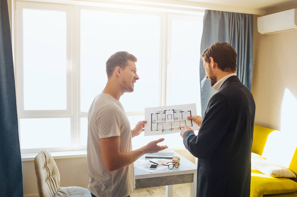 Young man and realtor buy or rent apartment. They hold one plan together. They talk and have business deal. Bright daylight comes out of window. - Foto, imagen