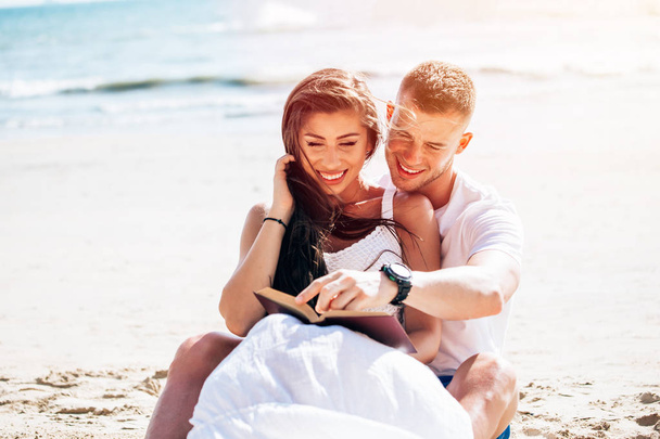Young happy and joyful Caucasian adult romantic couple in white summer wear - Boyfriend and girlfriend reading a book together on tropical beach - Photo, Image