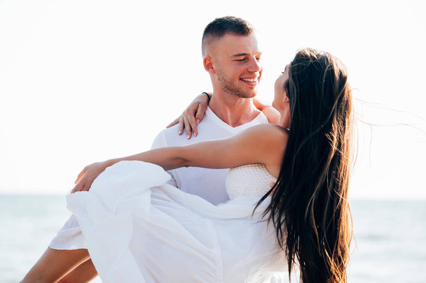 Young happy and joyful Caucasian adult romantic couple in white summer wear - Boyfriend carrying girlfriend on tropical beach and ocean - with copy space - 写真・画像