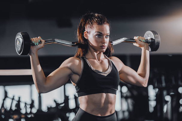 Attractive Caucasian female bodybuilder with ponytail lifting barbell while standing in gym. Train hard and do your best. - Photo, Image