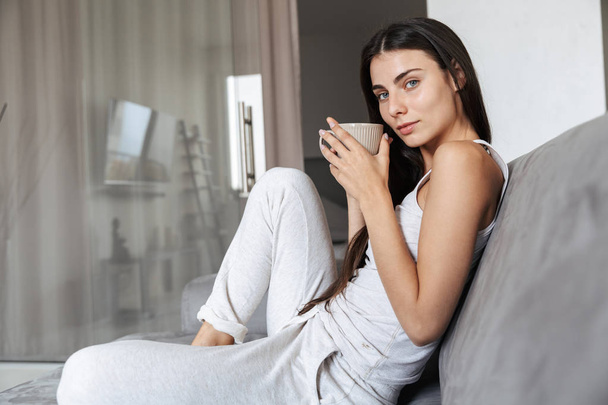 Pretty beautiful young woman sitting on sofa indoors at home drinking coffee or tea. - Photo, Image