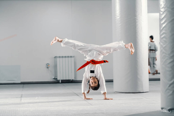 Young Caucasian girl in dobok doing hand stand with spread legs on taekwondo class. - Photo, Image