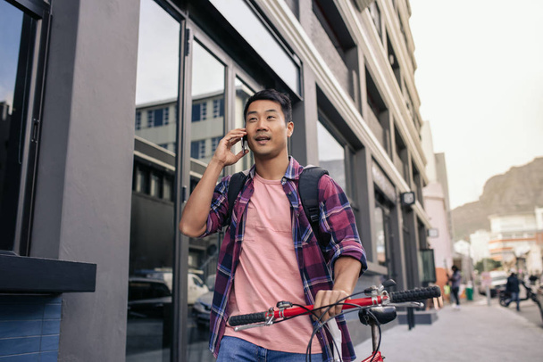 Young Asian man in a plaid shirt walking with his bicycle along a city sidewalk talking on a cellphone - Fotó, kép