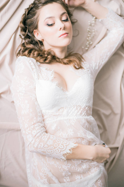 Young cute bride with beautiful hair in the morning at home in white lace negligee - Фото, изображение