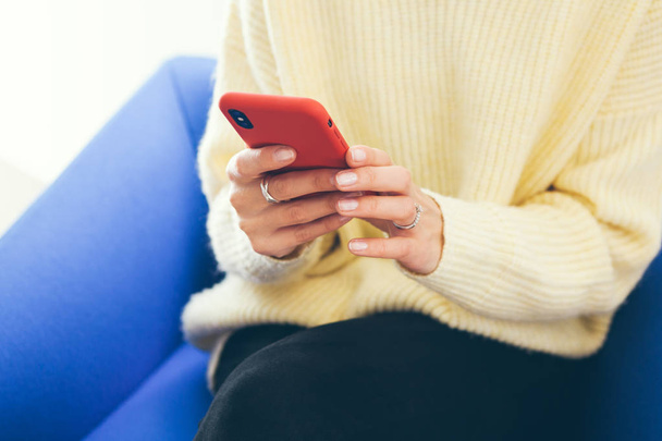 Lifestyle photo. Close-up of woman hands holding smartphone in red modern case and reading news, social network, surfing the web / internet in smartphone. Blue sofa background. - Foto, Imagem