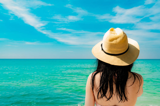 Πίσω όψη του σέξι και χαρούμενοι, νεαρή γυναίκα της Ασίας φορούν ψάθινο καπέλο χαλάρωση και απολαύστε διακοπές στην παραλία τροπικό παράδεισο. Κορίτσι στέκεται στην παραλία και κοιτάζοντας τη θάλασσα στις καλοκαιρινές τους διακοπές. Vibes καλοκαίρι. - Φωτογραφία, εικόνα