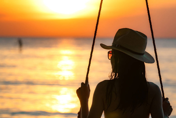 Sylwetka kobiety nosić bikini i słomkowy kapelusz huśtawka huśtawki na plaży na letnie wakacje o zachodzie słońca. Dziewczyny w strojach kąpielowych siedzieć na huśtawki i oglądać piękny zachód słońca. Letnie wibracje. Kobieta podróżować samotnie.  - Zdjęcie, obraz