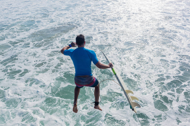 Surfista com prancha de surf salta do cais de praia molhe na água do oceano abaixo para entrada rápida para surfar ondas uma parte traseira close-up atrás para baixo foto de ação
. - Foto, Imagem