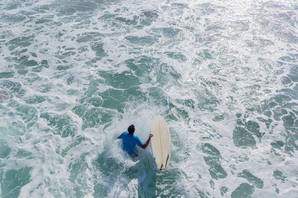 Surfer with surfboard jumps off beach pier jetty in ocean water below for quick entry out to surfing waves a closeup rear behind downward photo of action. - Foto, imagen