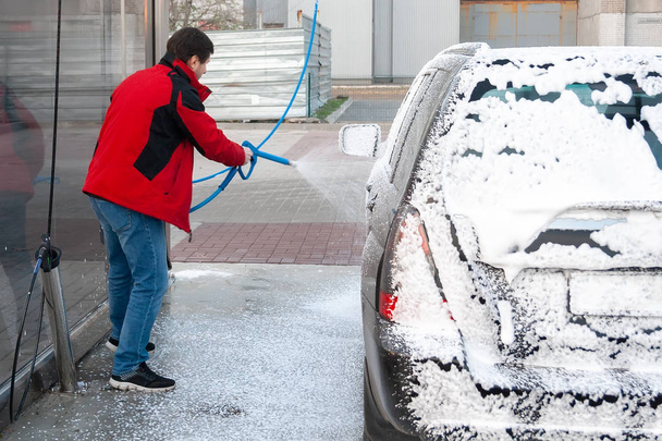 Un hombre con una chaqueta roja cubre un coche negro con espuma en un auto-servicio de lavado.
 - Foto, imagen