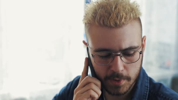 Potrait of young attractive businessman wearing glasses with yellow hair talking on the phone. Sitting at the desk with laptop. - Metraje, vídeo