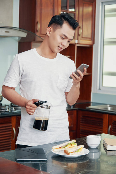 Casual Asian man holding pitcher with coffee and using smartphone in kitchen at home - Фото, изображение