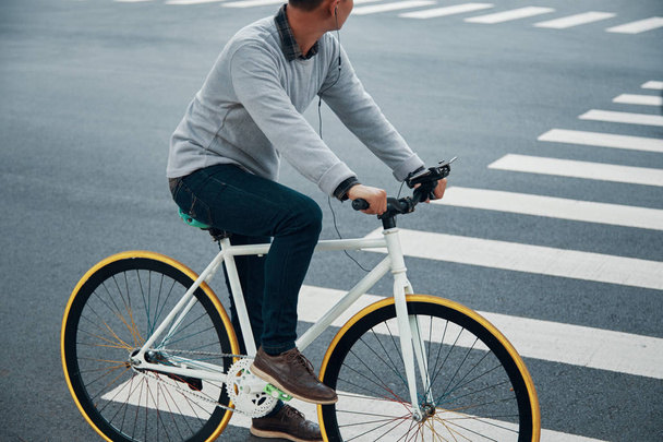 Seitenansicht eines anonymen Mannes, der Fahrrad entlang der Stadtstraße fährt und gute Musik hört - Foto, Bild