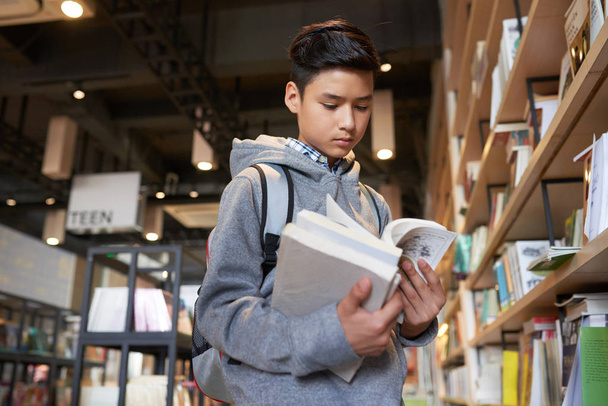 Joven adolescente con libros de celebración mirando a través de pie junto a las estanterías
 - Foto, imagen