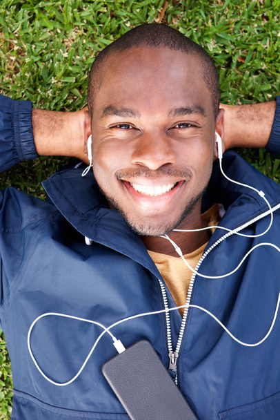 Portrait from above of smiling young black man lying on grass listening to music with earphones - Foto, Imagem