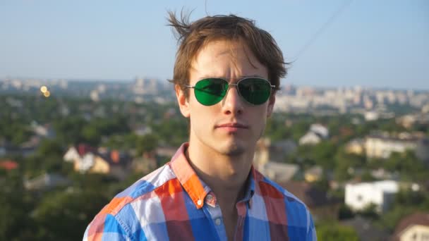 Close up portrait of young man in sunglasses sitting on rooftop of high-rise building with cityscape background. Handsome guy confident looks into camera with beautiful blurred landscape. Slow motion - Footage, Video