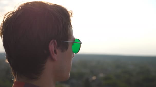 Das Profil eines jungen Mannes mit Sonnenbrille, der vom Dach eines Hochhauses das Stadtpanorama beobachtet. Der gutaussehende Kerl betrachtet das Stadtbild vom Dach aus und genießt die schöne Aussicht. Seitenansicht Nahaufnahme Zeitlupe - Filmmaterial, Video