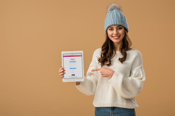 Blissful curly woman in knitted hat pointing with finger at digital tablet with instagram app on screen isolated on beige - Photo, image