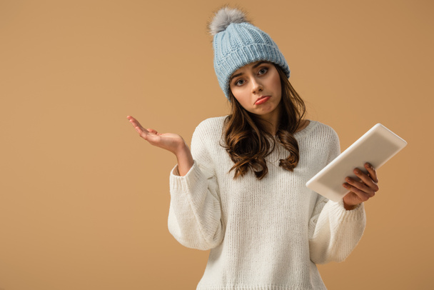 Giovane donna triste in cappello a maglia con tablet digitale isolato su beige
 - Foto, immagini