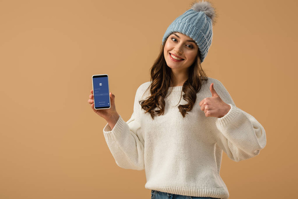 Mujer atractiva en sombrero de punto con teléfono inteligente con aplicación de Facebook en la pantalla y mostrando el pulgar hacia arriba aislado en beige
 - Foto, Imagen