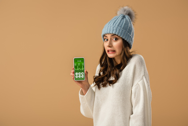 Confused curly young woman holding smartphone with health app on screen isolated on beige - Foto, afbeelding