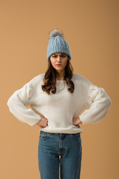 ベージュに分離された両手を腰に当て立っている白いセーターに懐疑的なブルネットの少女 - 写真・画像