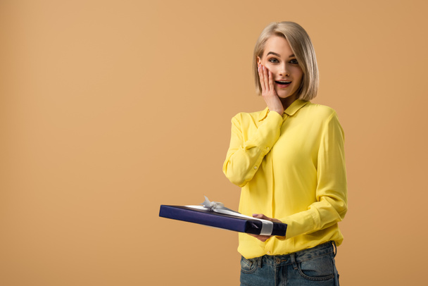 Surprised blonde woman in yellow shirt touching face and holding gift box isolated on beige - Photo, Image